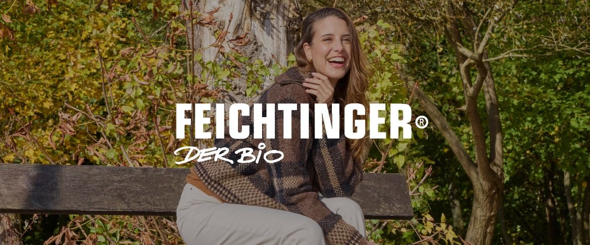 Eine Frau in einer karierten Jacke sitzt lächelnd auf einer Bank in einem herbstlichen Park. Das Logo der Marke Feichtinger Bio überlagert das Bild.