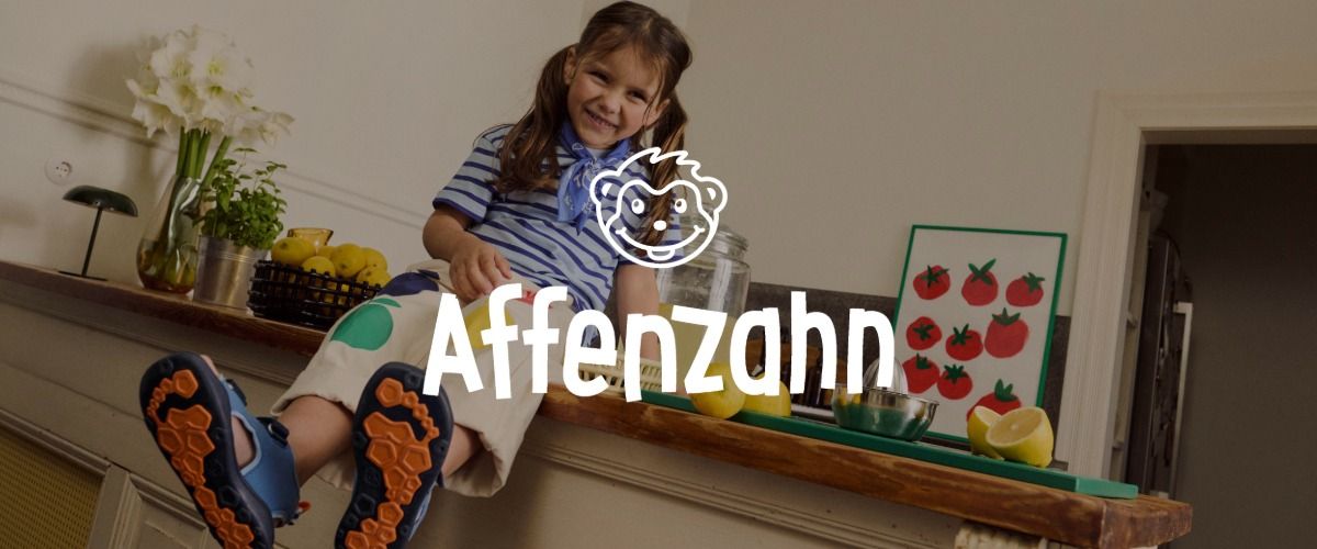 Ein lächelndes Kind mit Zöpfen sitzt in Freizeitkleidung auf einer Küchentheke, umgeben von Obst und einem Tomatenbild. Das Logo der Marke Affenzahn ist im Vordergrund.