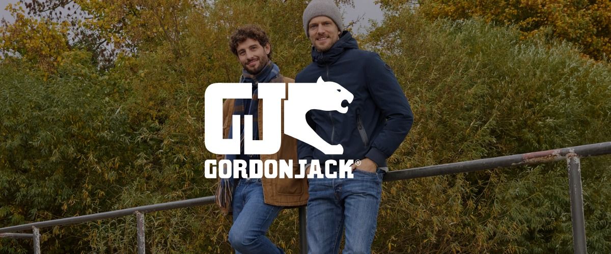 Zwei Männer posieren im Freien auf einem Metallgeländer, umgeben von üppigem Grün. Das Logo der Marke GORDON JACK überlagert das Bild.