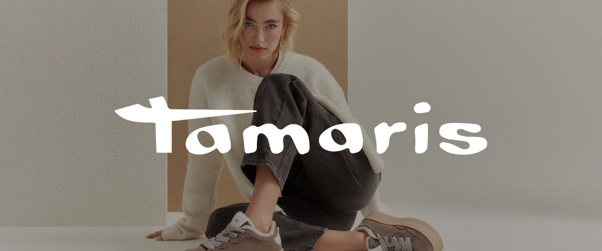 Eine Frau in weißem Strickpullover, die mit angewinkeltem Bein posiert und braune Sneaker trägt. Das Logo der Marke Tamaris überlagert das Bild.