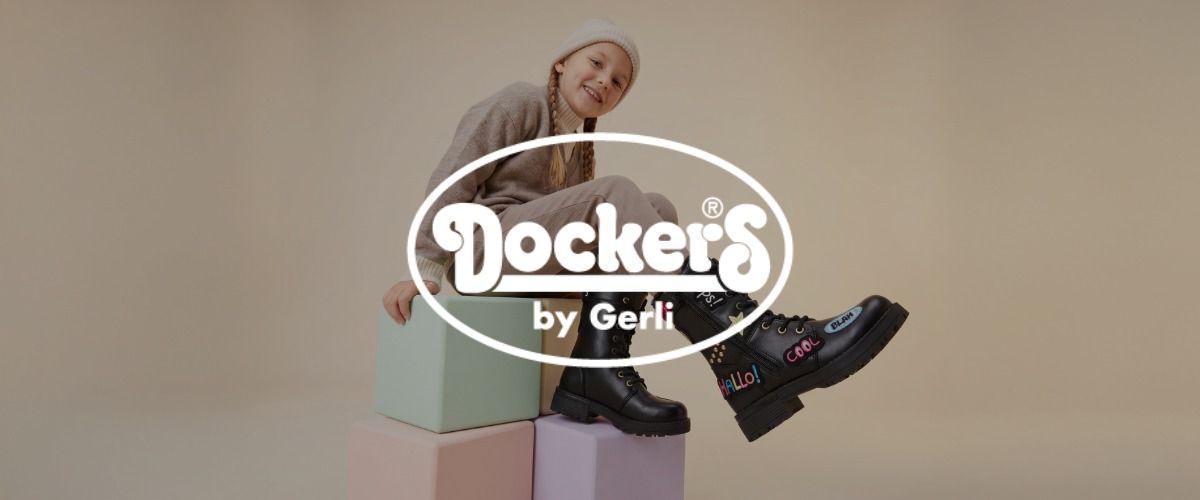 Ein Mädchen sitzt auf pastellfarbenen Würfeln und trägt schwarze Stiefel mit Aufnähern, vor einem neutralen Hintergrund. Das Logo der Marke Dockers überlagert das Bild.