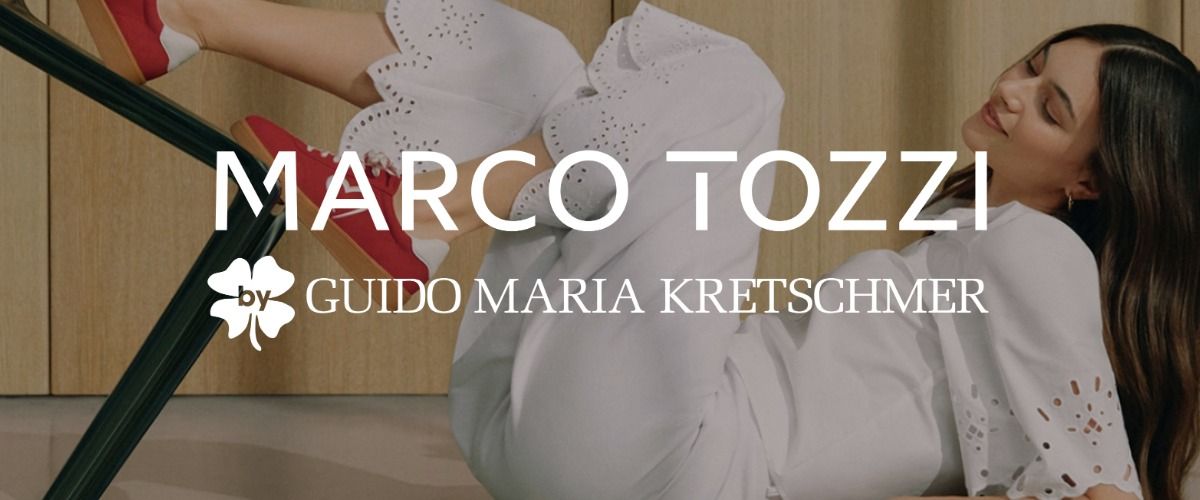 Eine Frau in einem weißen Outfit mit Lochstickerei liegt auf dem Boden mit den Beinen auf einem Stuhl. Sie trägt rote Sneaker. Das Bild wird von dem Logo der Marke Marco Tozzi by Guido Maria Kretschmer überlagert.