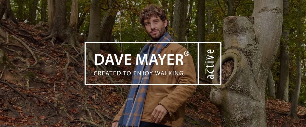 Ein Mann in einer braunen Jacke und einem blauen Schal steht in einem Wald. Das Logo der Marke DAVE MAYER active überlagert das Bild.