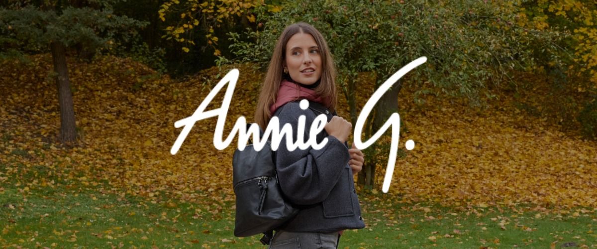 Eine Frau steht mit einem Rucksack in einem Park, umgeben von Herbstblättern und Bäumen. Das Logo der Marke Annie G. überlagert das Bild.