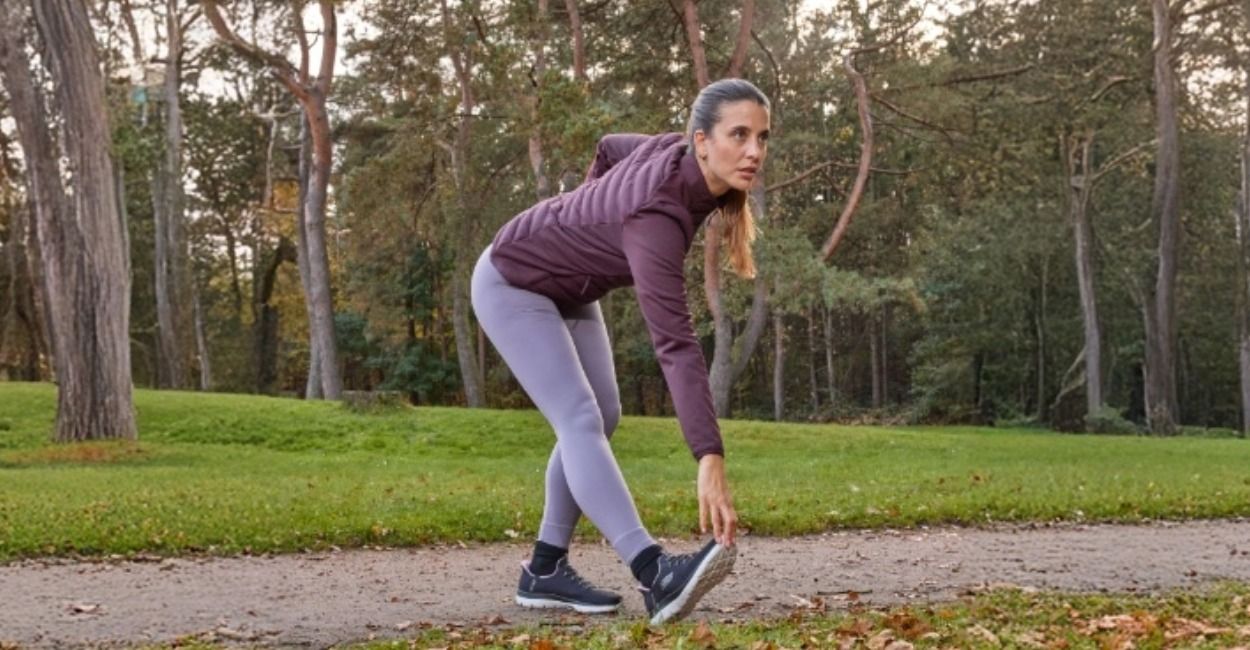 Eine Frau in Sportkleidung macht draußen in einem Park Dehnübungen und berührt auf einem von Bäumen gesäumten Feldweg ihre Zehen.