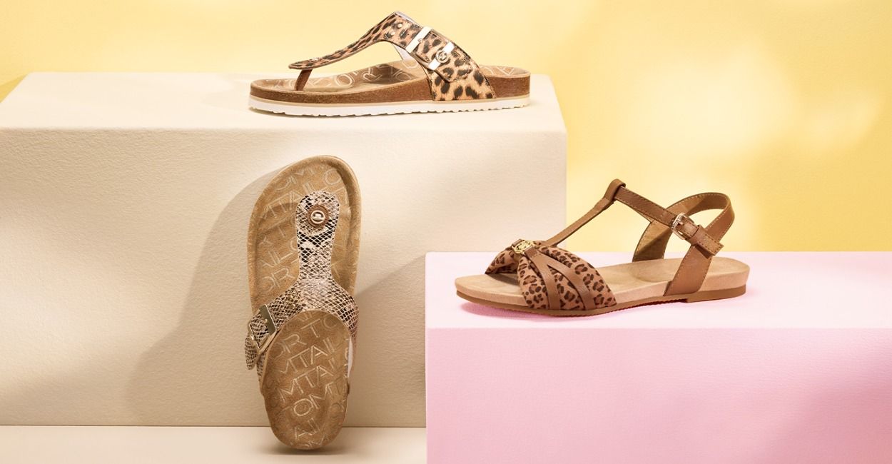 Zwei Sandalen mit Leopardenmuster und eine mit Schlangenmuster, präsentiert auf geometrischen Blöcken vor einem pastellgelben Hintergrund.