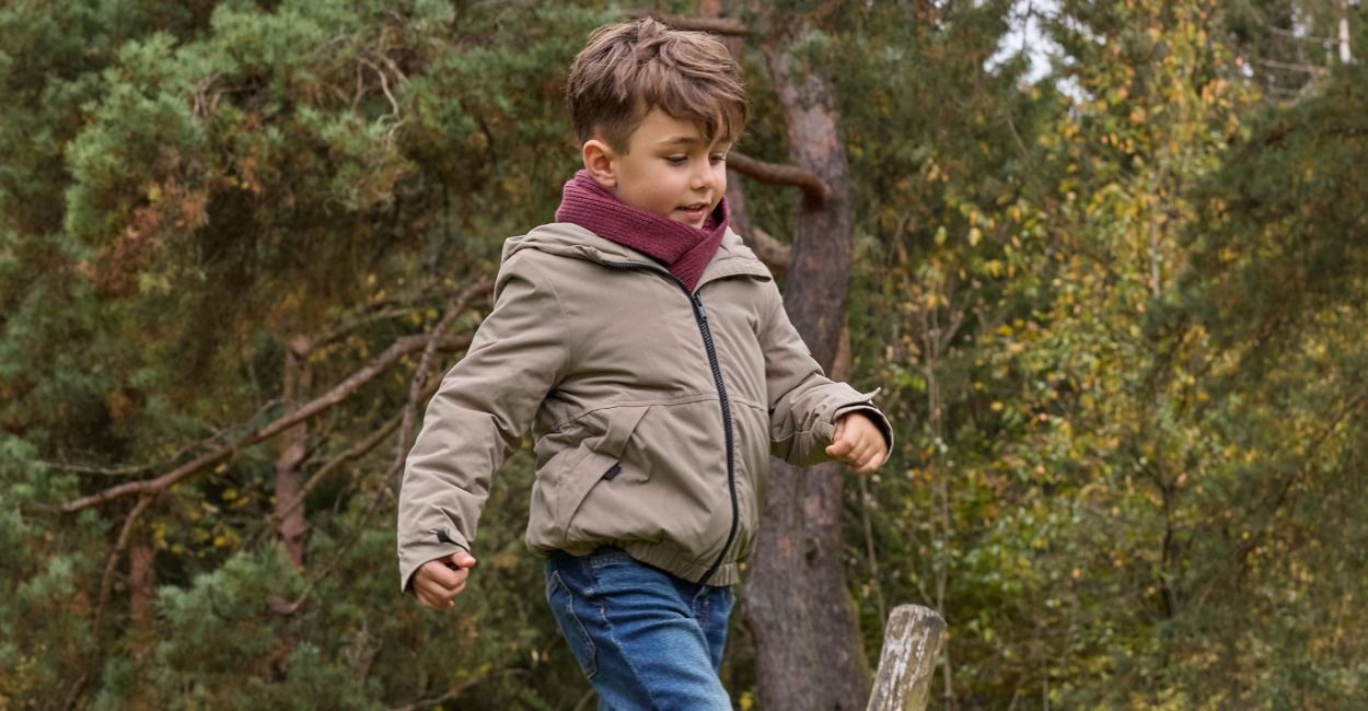 Ein kleiner Junge, der eine Jacke und einen Schal trägt, rennt fröhlich im Freien herum, umgeben von üppig grünen Bäumen.