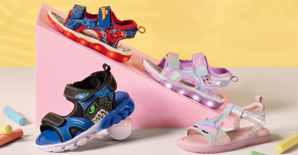 Vier farbenfrohe Kindersandalen mit Comic-Motiven vor einem gelben Hintergrund.