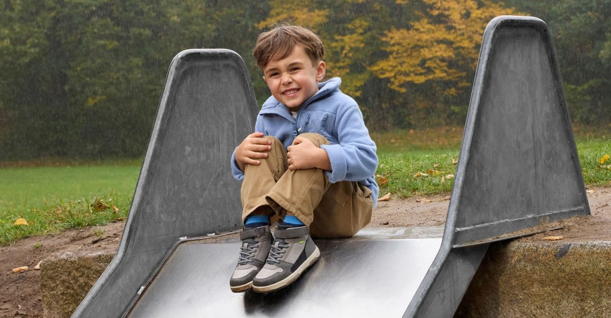 Ein kleiner Junge in einer blauen Jacke und einer beigen Hose sitzt an einem regnerischen Tag oben auf einer Rutsche in einem Park und lächelt, im Hintergrund sind herbstlich gefärbte Bäume zu sehen.