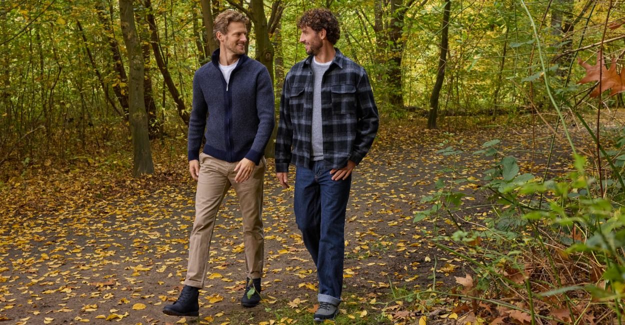 Zwei Männer, die im Herbst auf einem mit Laub bedeckten Waldweg spazieren gehen und sich dabei anlächeln.