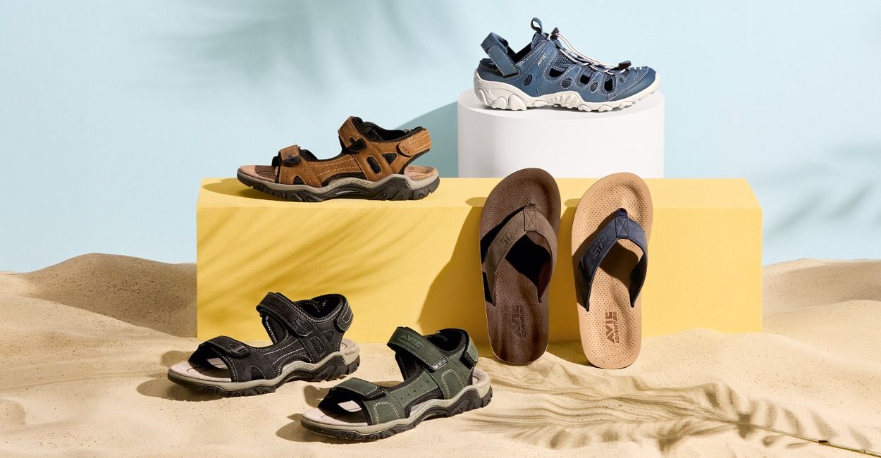 Auf Sand mit gelbem Podest sind verschiedene Sandalen ausgestellt: braune, grüne und blaue Wandersandalen sowie braune und blaue Flip-Flops.