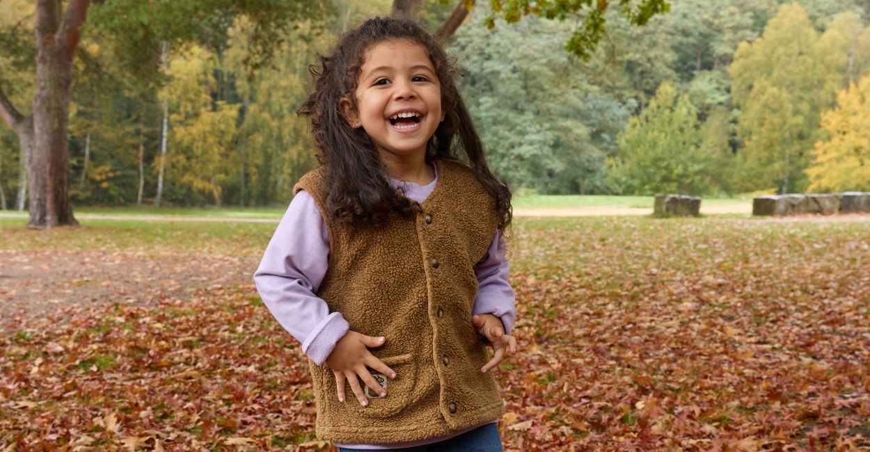 Ein fröhliches MÀdchen in einer braunen Weste und einem lila Pullover steht auf dem mit HerbstblÀttern bedeckten Boden eines Parks und lÀchelt breit, wÀhrend im Hintergrund BÀume zu sehen sind.