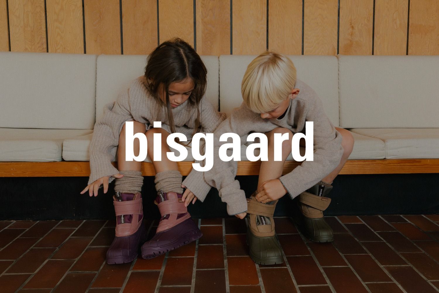 Zwei Kinder sitzen in Winterstiefeln auf einer Bank. Das Logo der Marke bisgaard überlagert das Bild.