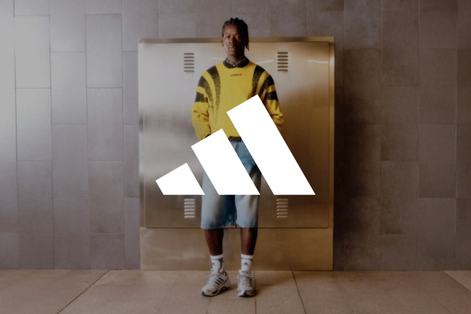 Eine Person in einem gelb-schwarzen Pullover, Jeansshorts und Turnschuhen steht vor einem metallischen Hintergrund. Das Logo der Marke adidas überlagert das Bild.
