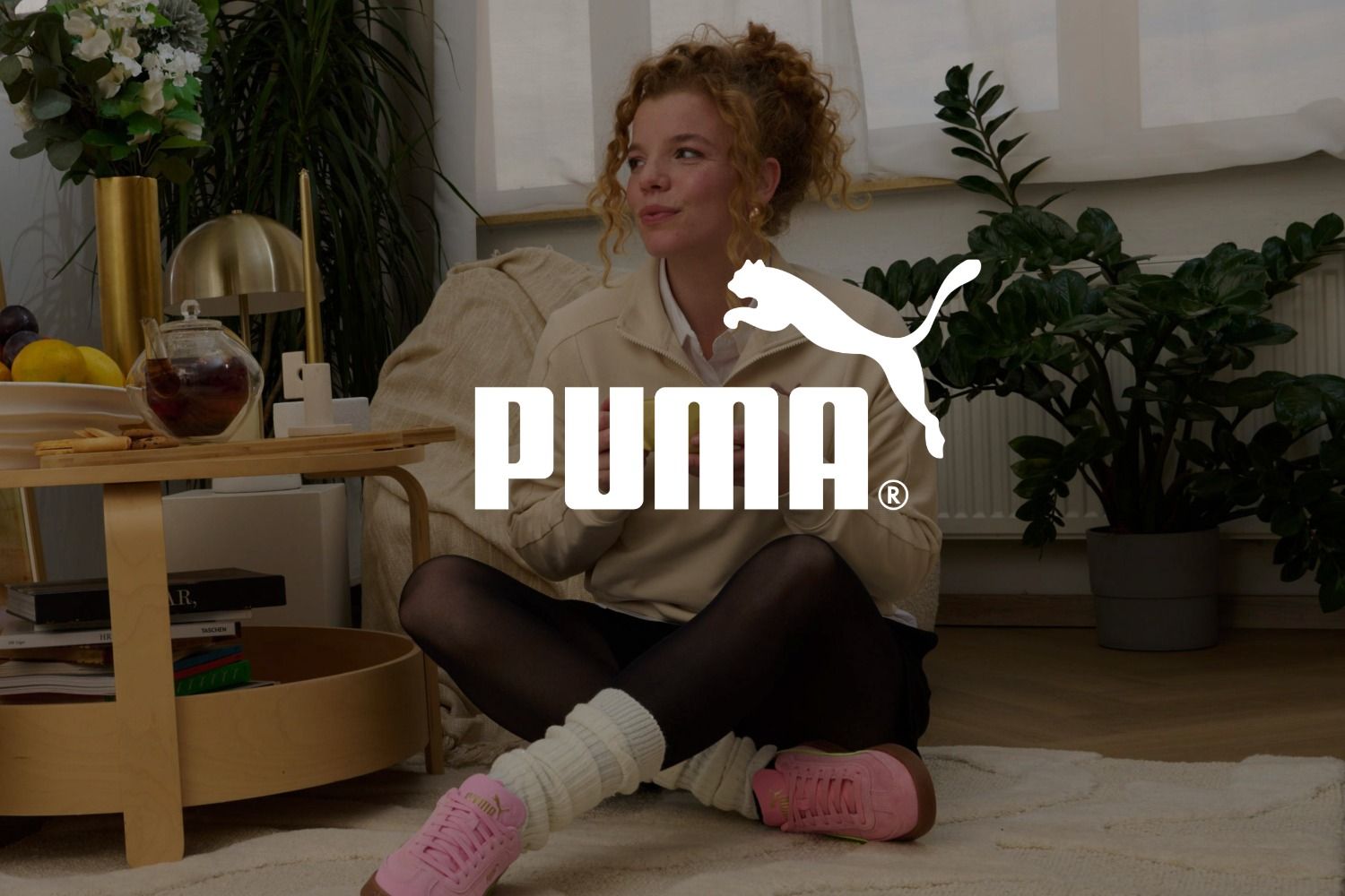 Eine Frau mit lockigem Haar sitzt auf dem Boden, trägt rosa Puma-Sneaker und eine schwarze Strumpfhose; im Hintergrund sind Pflanzen und ein Holztisch zu sehen. Das Logo der Marke PUMA überlagert das Bild.
