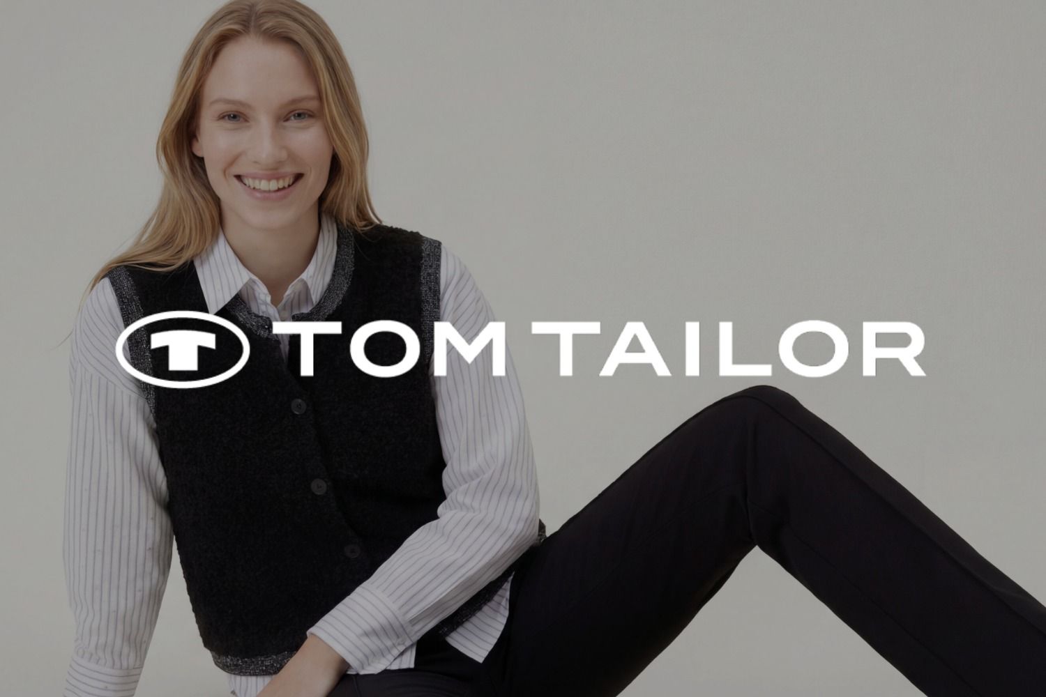 Eine lächelnde Frau, die ein gestreiftes Shirt und eine schwarze Weste trägt. Das Logo der Marke TOM TAILOR überlagert das Bild.