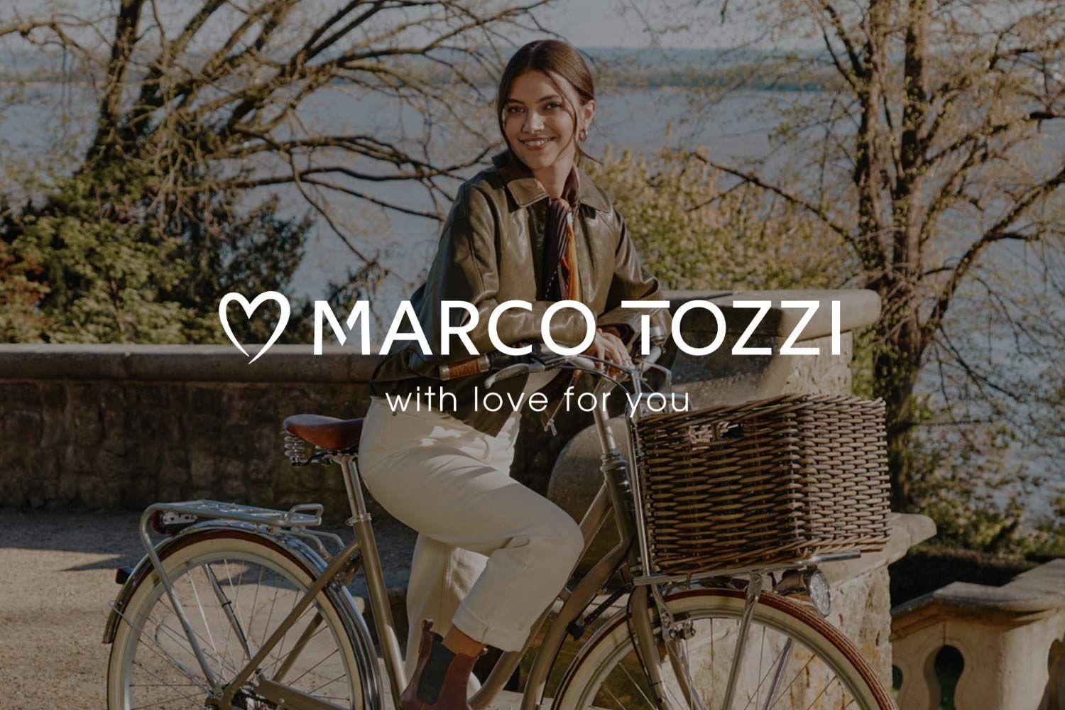 Eine Frau in einer grünen Jacke sitzt lächelnd auf einem Fahrrad mit einem Weidenkorb, im Hintergrund sind Bäume und ein Gewässer zu sehen. Das Logo der Marke MARCO TOZZI überlagert das Bild.