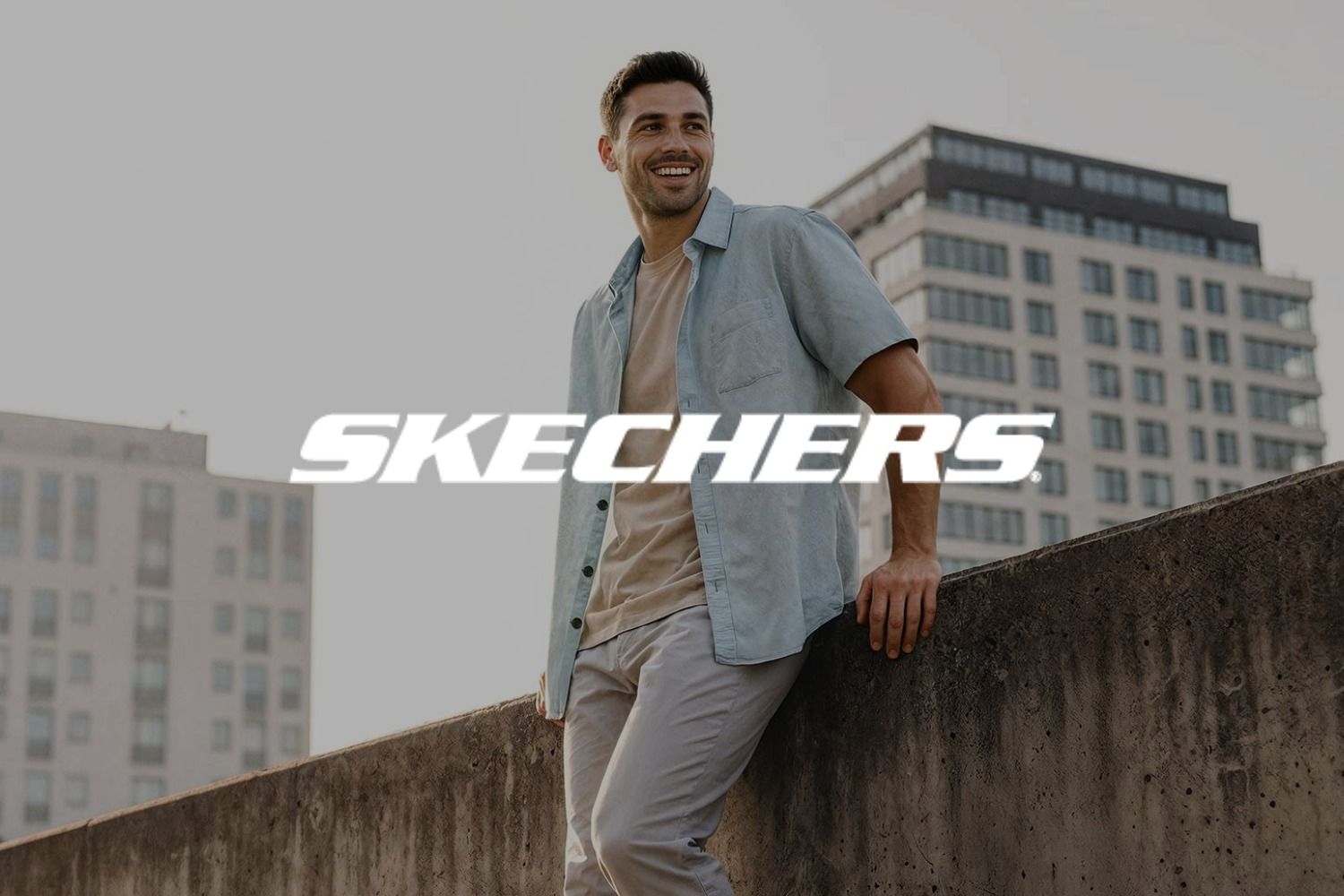 Ein Mann, der an einer Betonmauer lehnt, lächelt und Freizeitkleidung trägt. Im Hintergrund sind Gebäude vor einem klaren Himmel zu sehen. Das Logo der Marke Skechers überlagert das Bild.