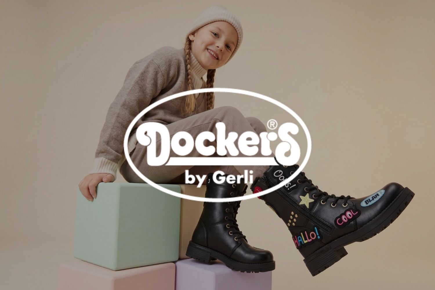 Ein lächelndes Mädchen in gemütlicher Kleidung sitzt auf bunten Bauklötzen und trägt schwarze Schnürstiefel mit verspielten Aufnähern. Das Logo der Marke Dockers überlagert das Bild.