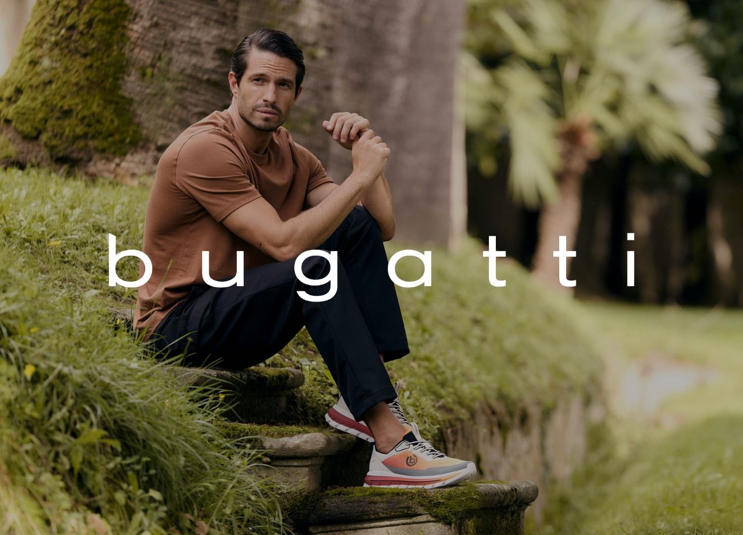 Mann sitzt auf Steinstufen in einem Garten, trägt ein braunes Hemd und Turnschuhe. Das Logo der Marke bugatti ist im Vordergrund.