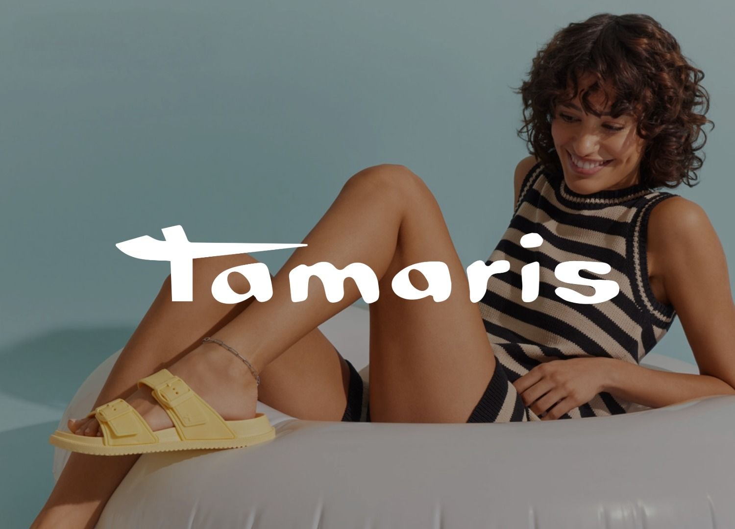 Eine lächelnde Frau in einem gestreiften Outfit und gelben Sandalen sitzt auf einem aufblasbaren Stuhl. Das Logo der Marke Tamaris überlagert das Bild.
