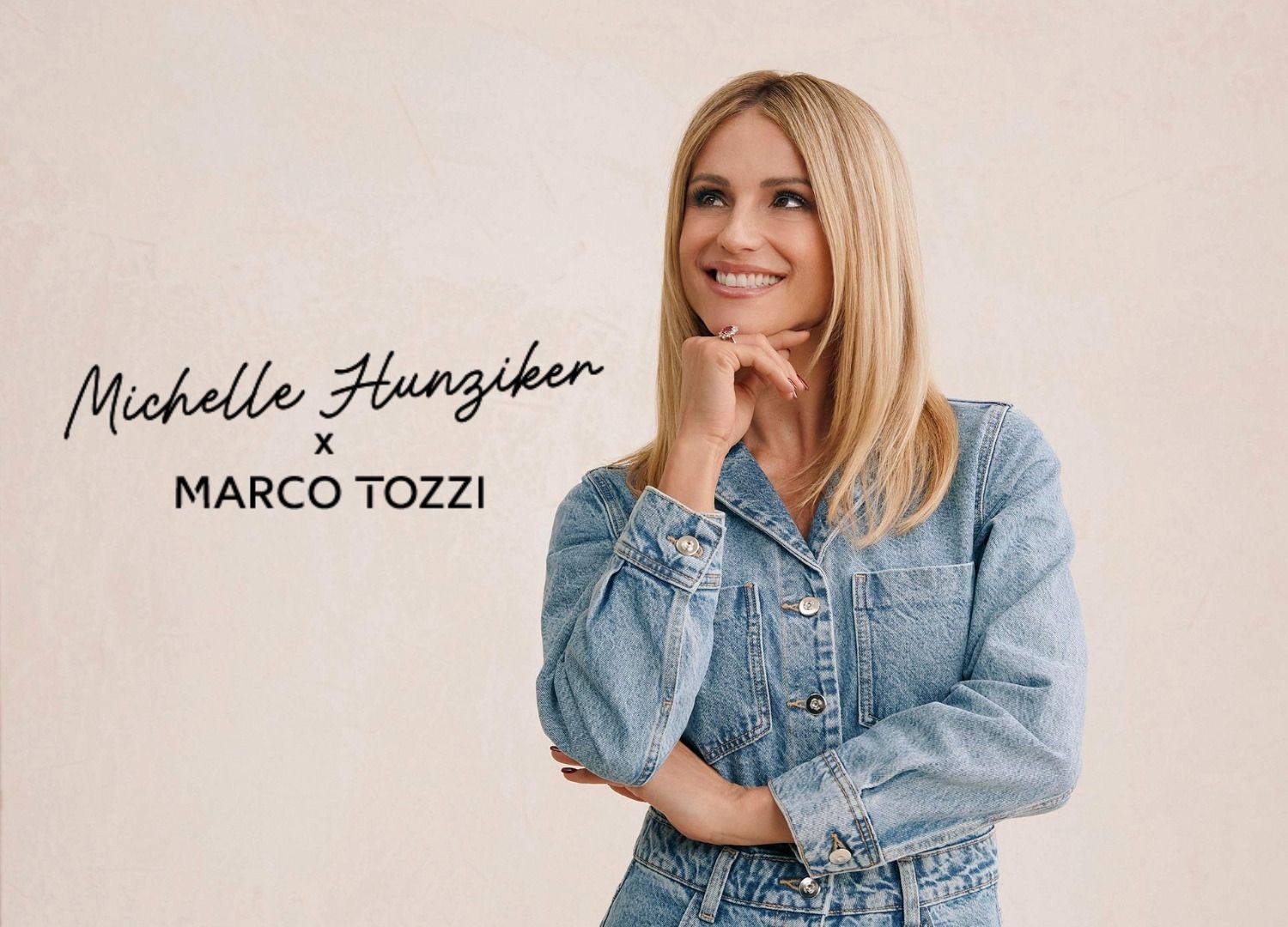 Eine Frau mit blonden Haaren, die einen Jeansoverall trägt und nachdenklich lächelt, während sie ihre Hand an das Kinn legt. Sie steht vor einem hellen Hintergrund.