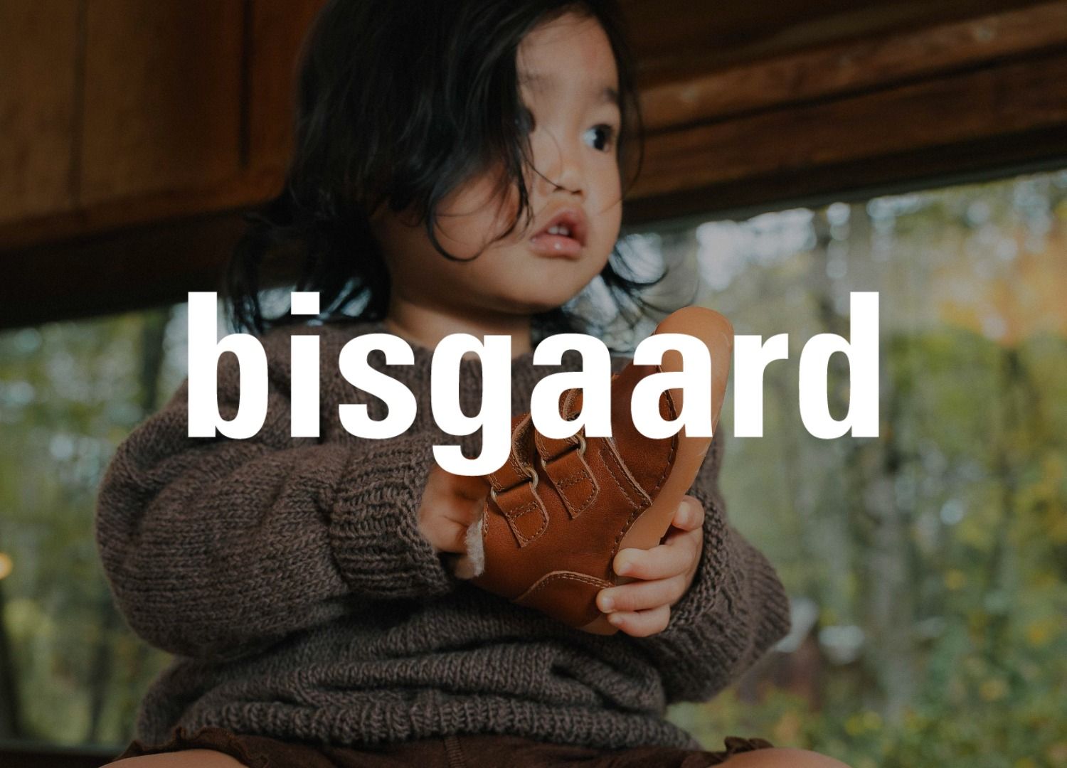 Ein Kind hält einen braunen Schuh in einem Raum mit Holzvertäfelung mit großem Fenster in der Hand. Das Logo der Marke bisgaard überlagert das Bild.