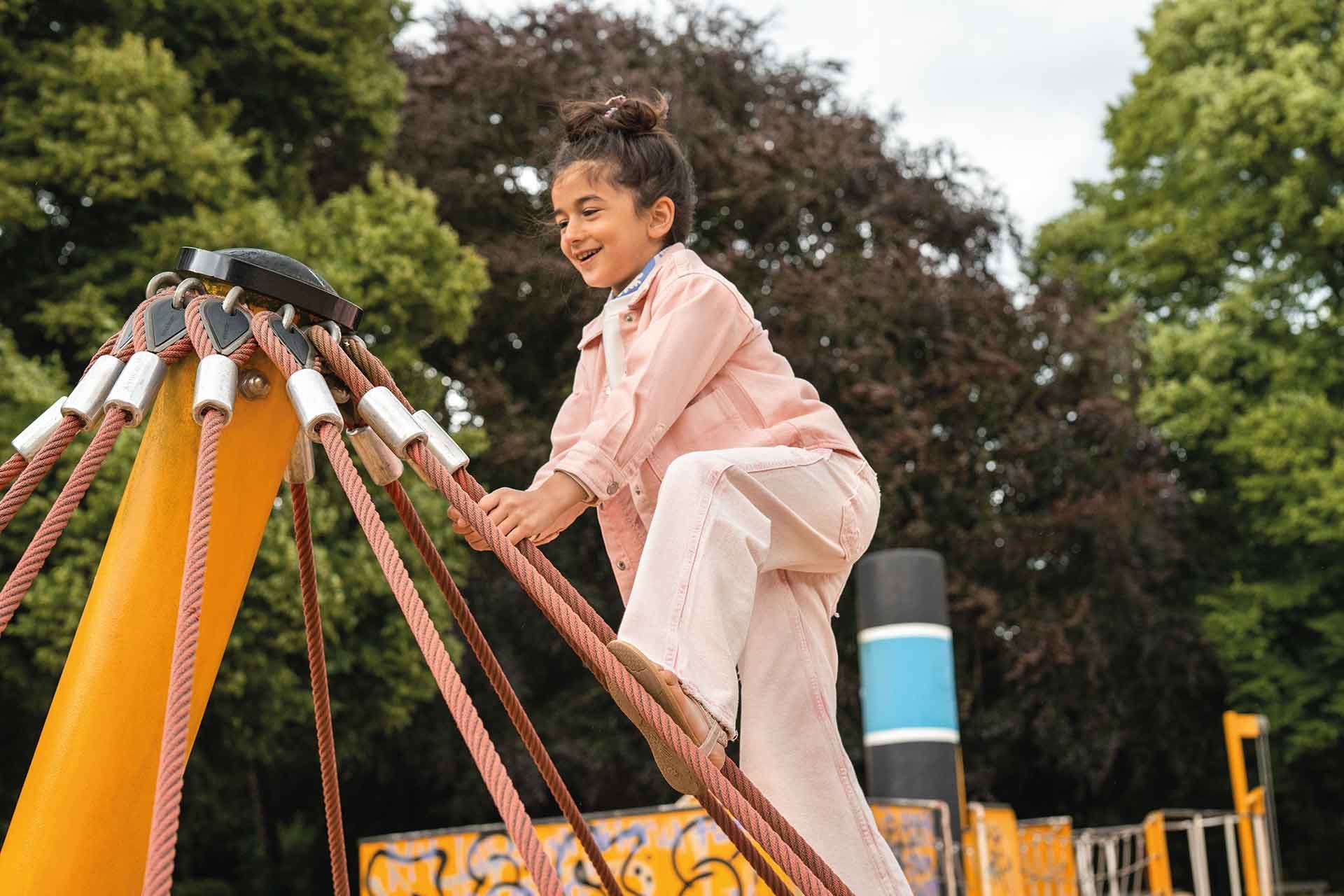 Ein junges Mädchen in einem rosa Outfit klettert auf einem Spielplatz, umgeben von Bäumen, auf eine bunte Seilkonstruktion.