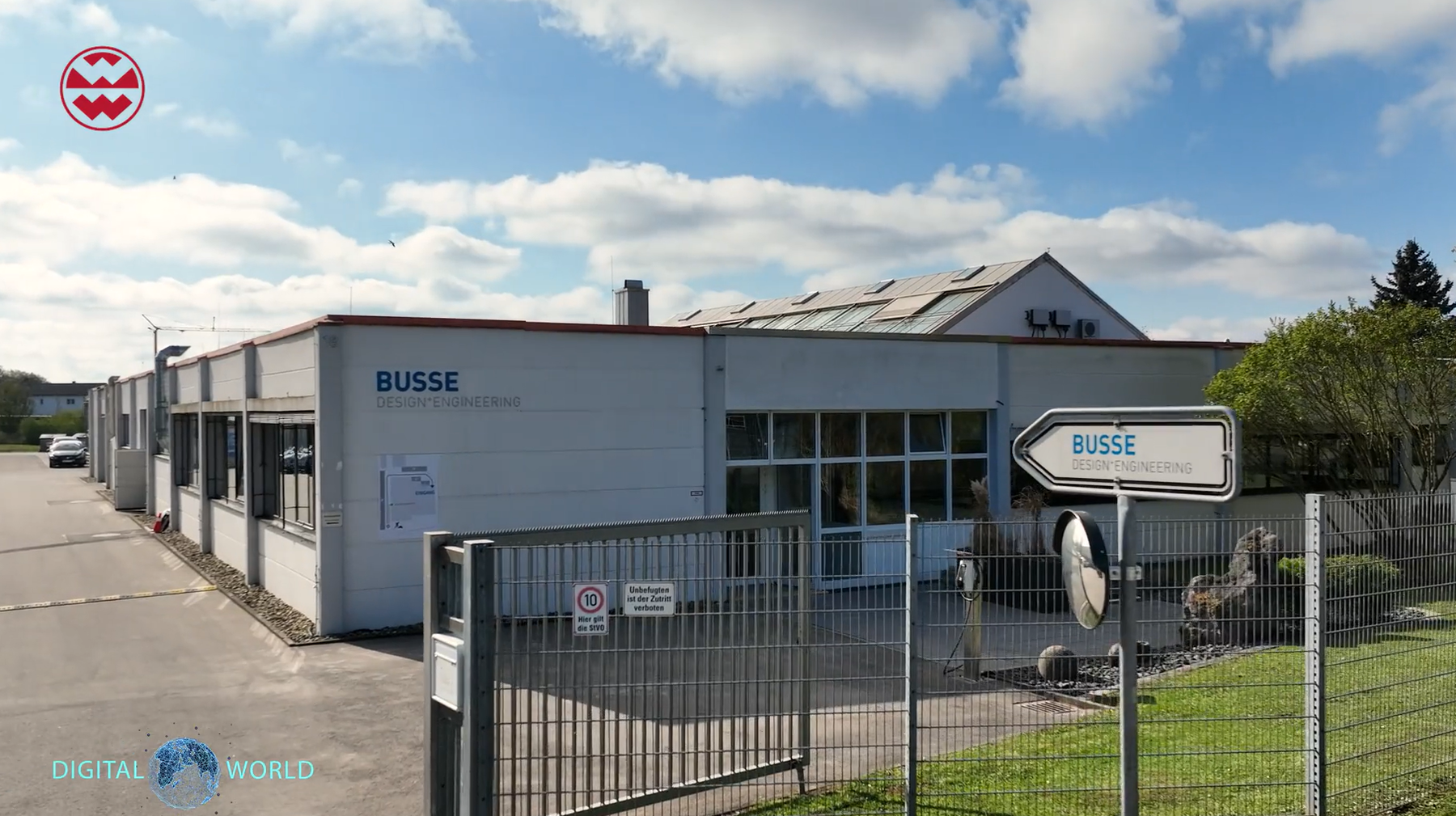Außenansicht des Gebäudes von Busse Design Engineering mit einem Zaun und einem Wegweiser unter teilweise bewölktem Himmel.