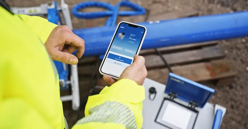 A person is standing at a building site, holding a mobile phone with the Georg Fischer app open.