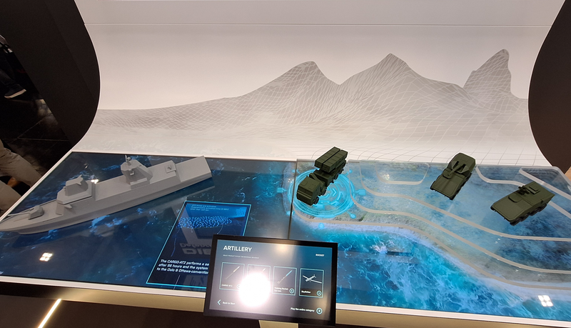 Ein Bildschirm, auf dem eine Meeres- und Landlandschaft zu sehen ist, auf der ein Modellschiff und mehrere Modellfahrzeuge platziert sind.