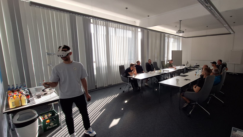Eine Person mit einem VR-Headset steht in einem sonnendurchfluteten Büro, während mehrere Personen an Tischen sitzen, Getränke und Snacks zu sich nehmen und beobachten.