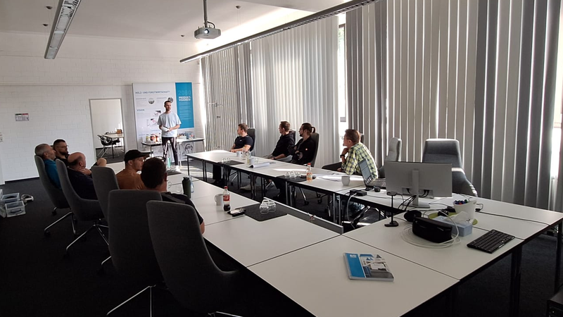 Eine Gruppe von Personen sitzt an Tischen in einem Konferenzraum und hört einem Vortragenden zu, der neben einer Leinwand steht.