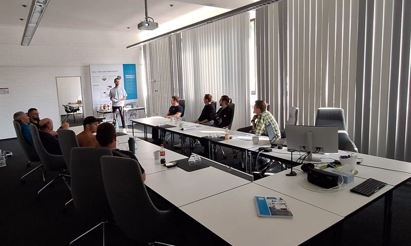 Eine Gruppe von Personen sitzt an Tischen in einem Konferenzraum und hört einem Vortragenden zu, der neben einer Leinwand steht.