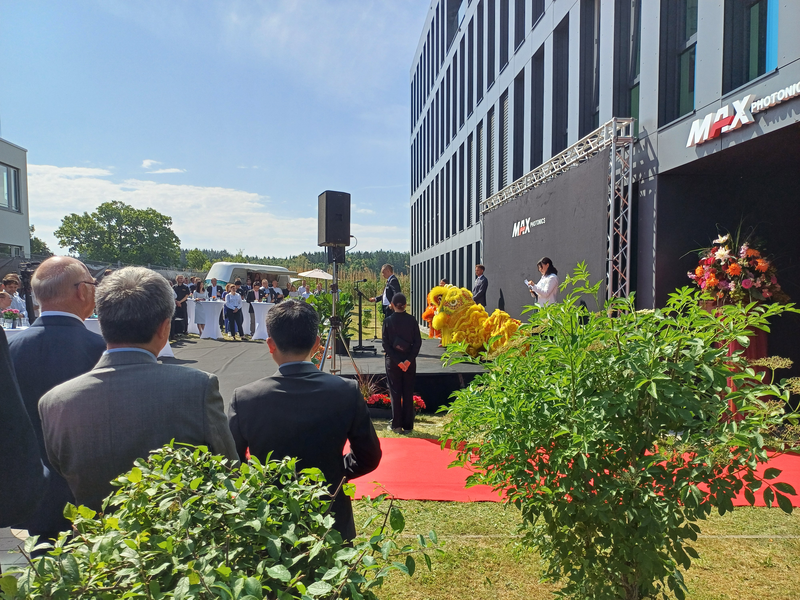 A lecture is being held on a stage in front of the Maxphotonics building.