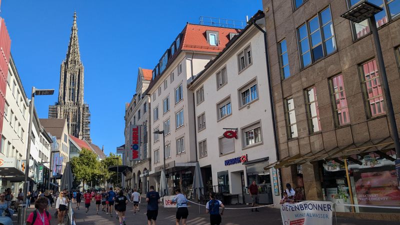 The marathon runners are jogging along a shopping street, with Ulm Minster visible in the background.