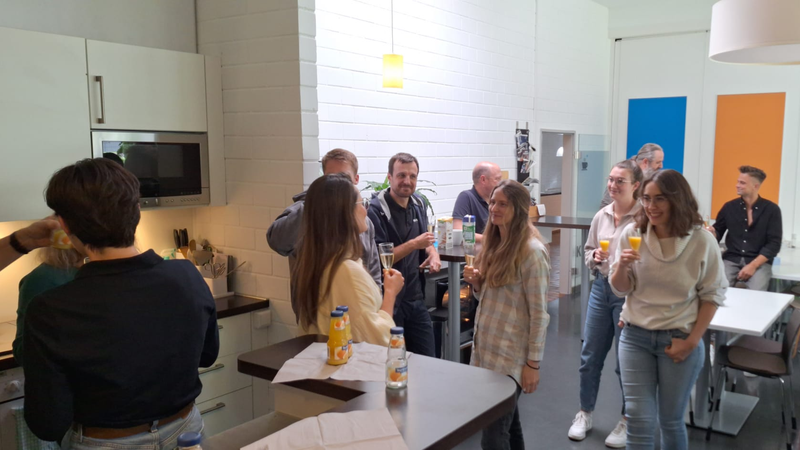 Eine Gruppe von Menschen in einem Küchenbereich, die sich unterhalten und Getränke in der Hand halten, in einer Mischung aus legerer Kleidung und modernem Ambiente.