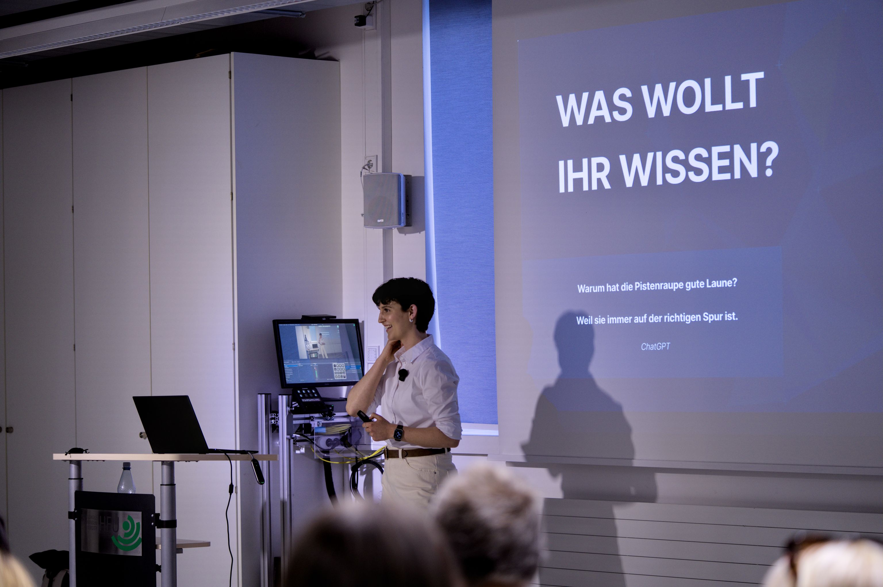 A person is giving a presentation in a seminar room in front of a screen; a laptop and a monitor are standing next to it.
