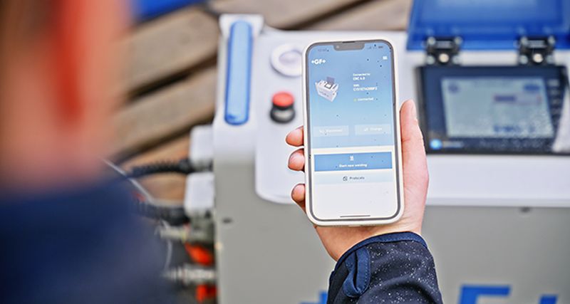 Person holding a smartphone displaying an app interface near electronic equipment.