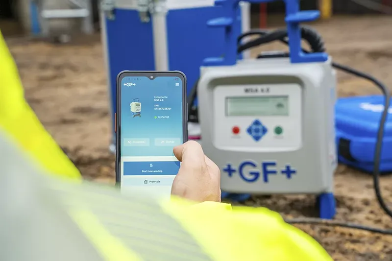 Person using a smartphone app to connect with a welding machine marked "+GF+" on-site, wearing a yellow safety jacket.