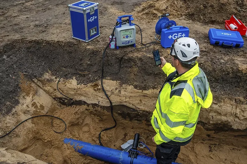 A worker wearing a bright yellow jacket and a hard hat is laying underground pipes on a building site with the aid of machinery.