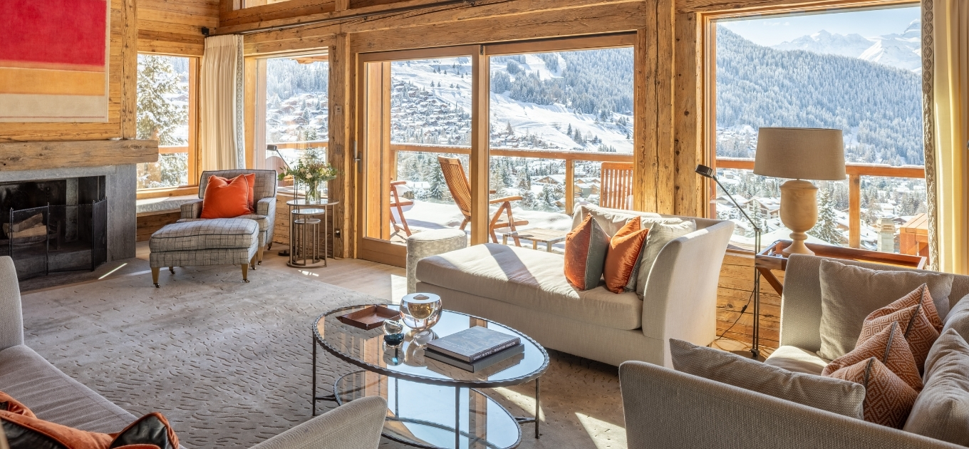 Cozy living room with a fireplace, soft couches, and large windows offering a scenic view of snow-covered mountains and forests in the background.