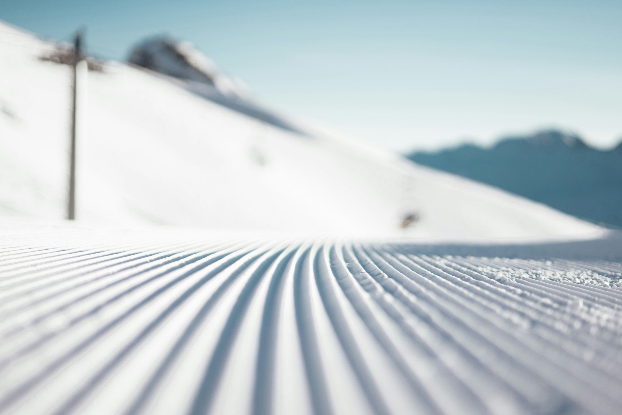 Groomed ski slope with parallel grooves under a clear blue sky, mountains in the background.
