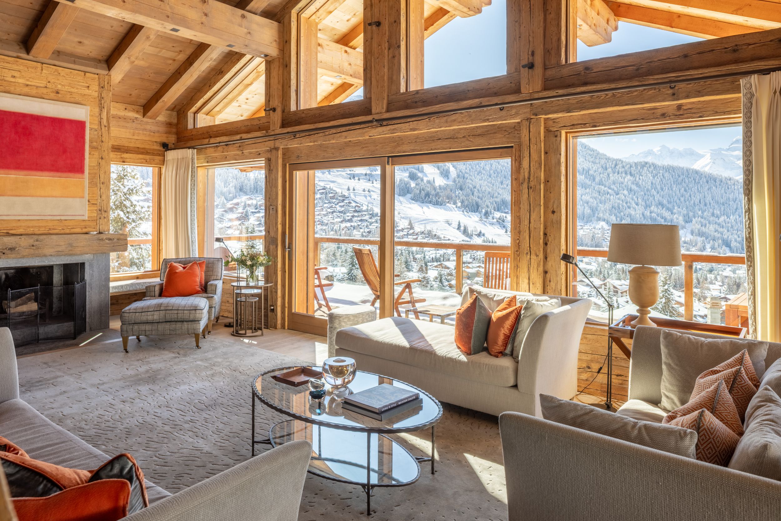 Cozy chalet living room with wooden beams, large windows offering a snowy mountain view, white and orange decor, fireplace, and plush seating.