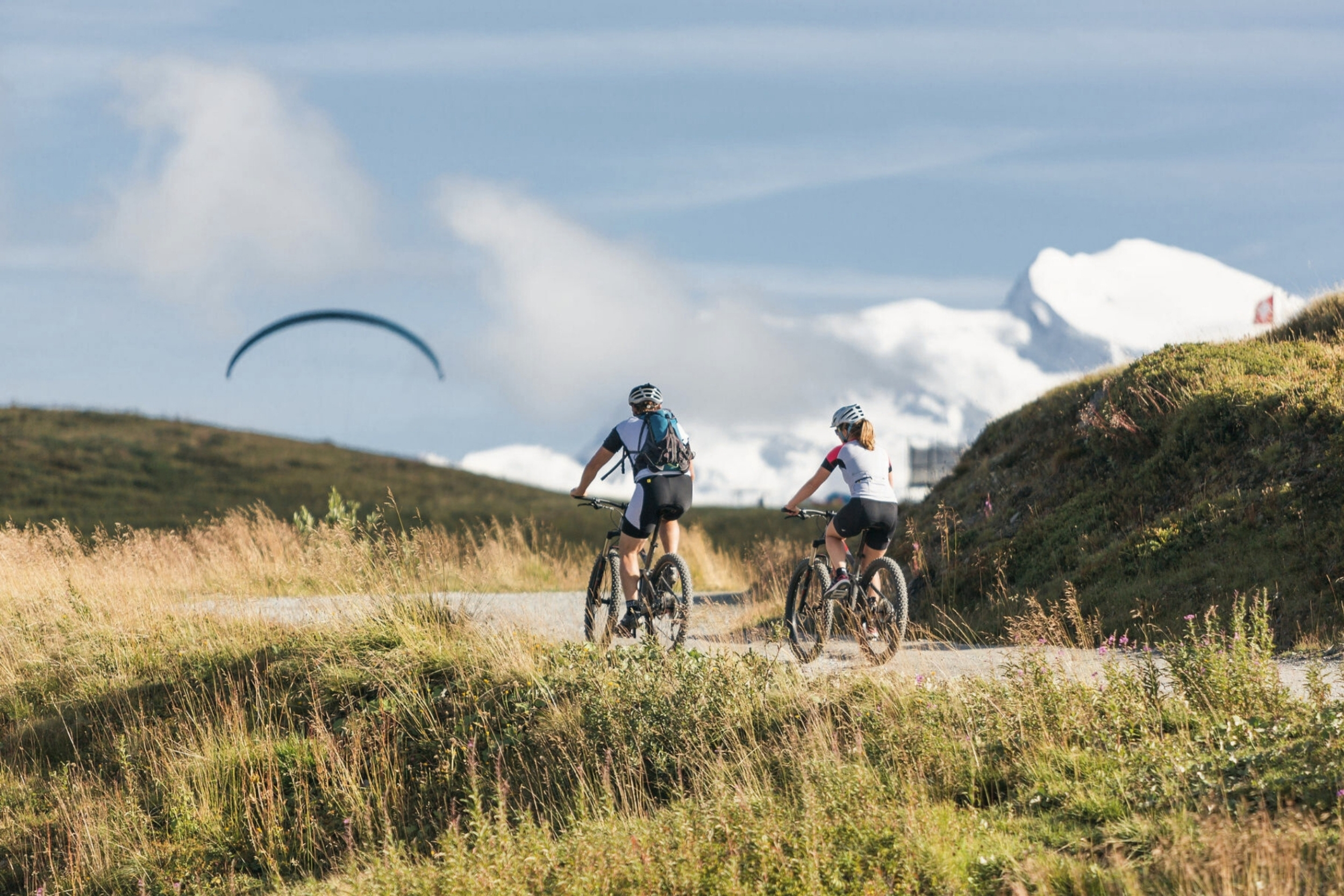 two bikers on a mountain trail in verbier in summer with a paraglider and snowy peaks in the background