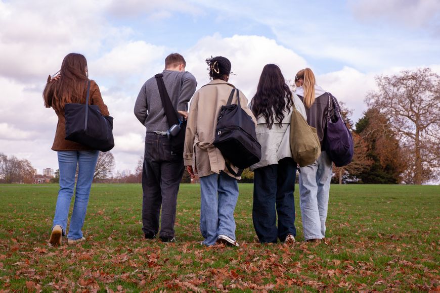 Five people walking on a grassy field with scattered leaves, carrying bags, under a partly cloudy sky.