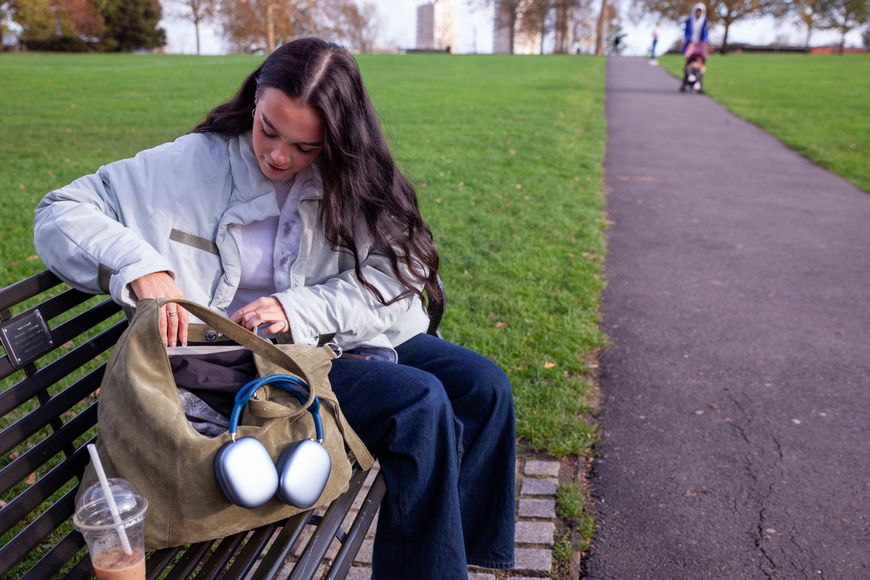 A woman sitting on a park bench rummaging through a bag with headphones hanging from it. A drink is beside her, and a stroller is in the background.