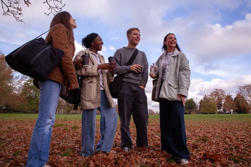 Four people stand and chat in a park on a cloudy day, surrounded by fallen leaves. They appear relaxed and carry bags.