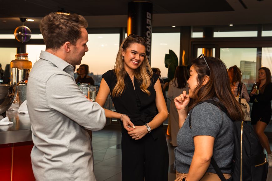 three people standing around a bar with the sunset in the background chatting, the person on the left has a backpack on, the person in the middle is wearing black and the person on the right is wearing a grey shirt