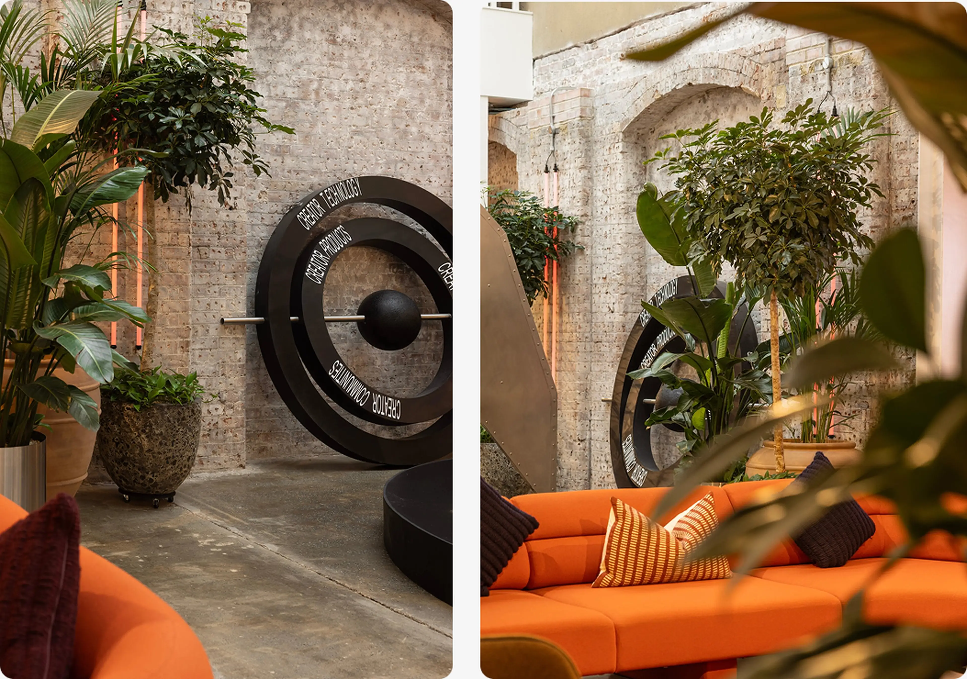 Cozy indoor lounge with orange sofa, decorative pillows, lush green plants, and a large artistic metal sculpture against a brick wall backdrop.
